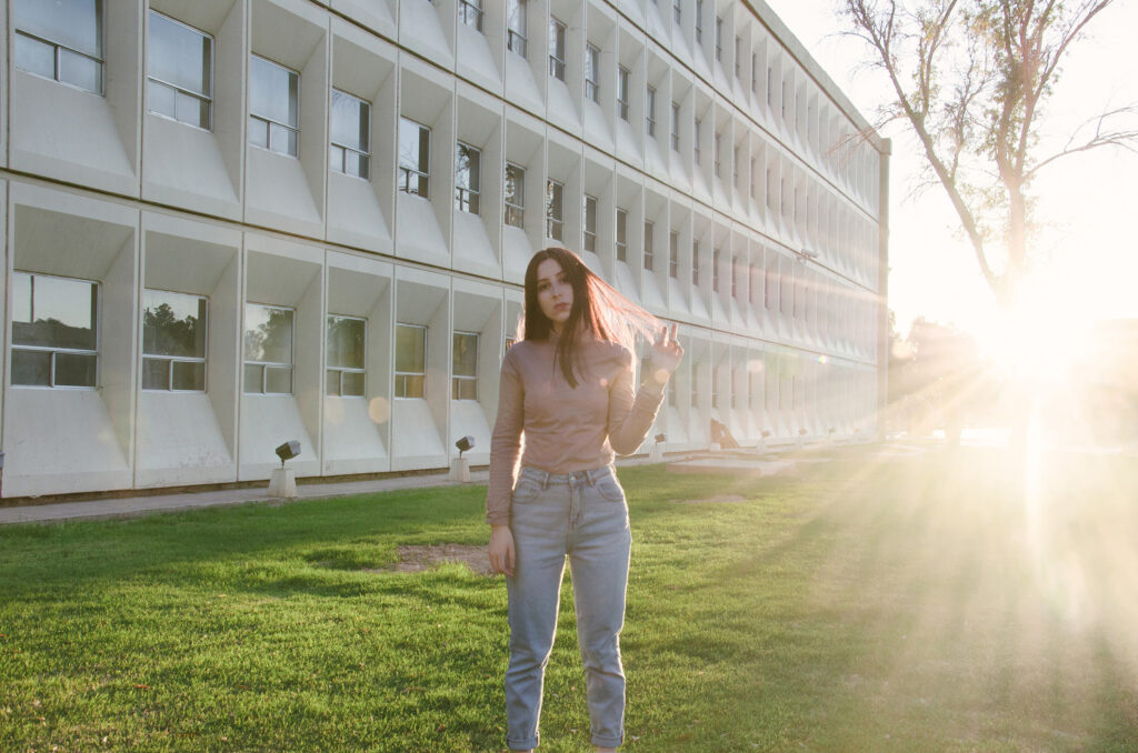 Gabriela Inzunza stands in front of a modern building shielding her eyes from the sun's glare with the golden hour light creating a radiant ambiance.